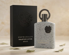 A bottle of Afnan Supremacy Collector's Edition fragrance stands beside its black presentation box on a light stone surface with scattered leaves. The bottle has a speckled light gray finish and a dark metallic ring cap, while the box is matte black with embossed branding.